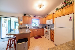 A kitchen with wooden cabinets, a white refrigerator, a microwave, and a stove. There is a window with blinds and a door leading to another room.