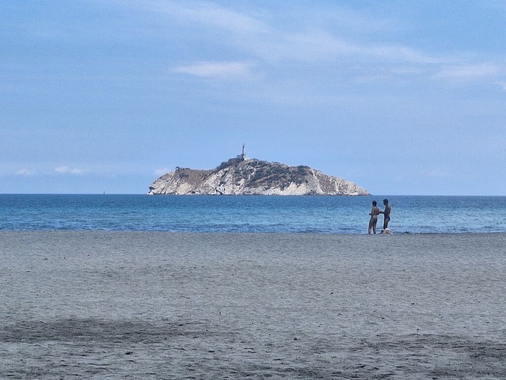 Une plage avec une île rocheuse à l’horizon et deux personnes debout sur le sable.