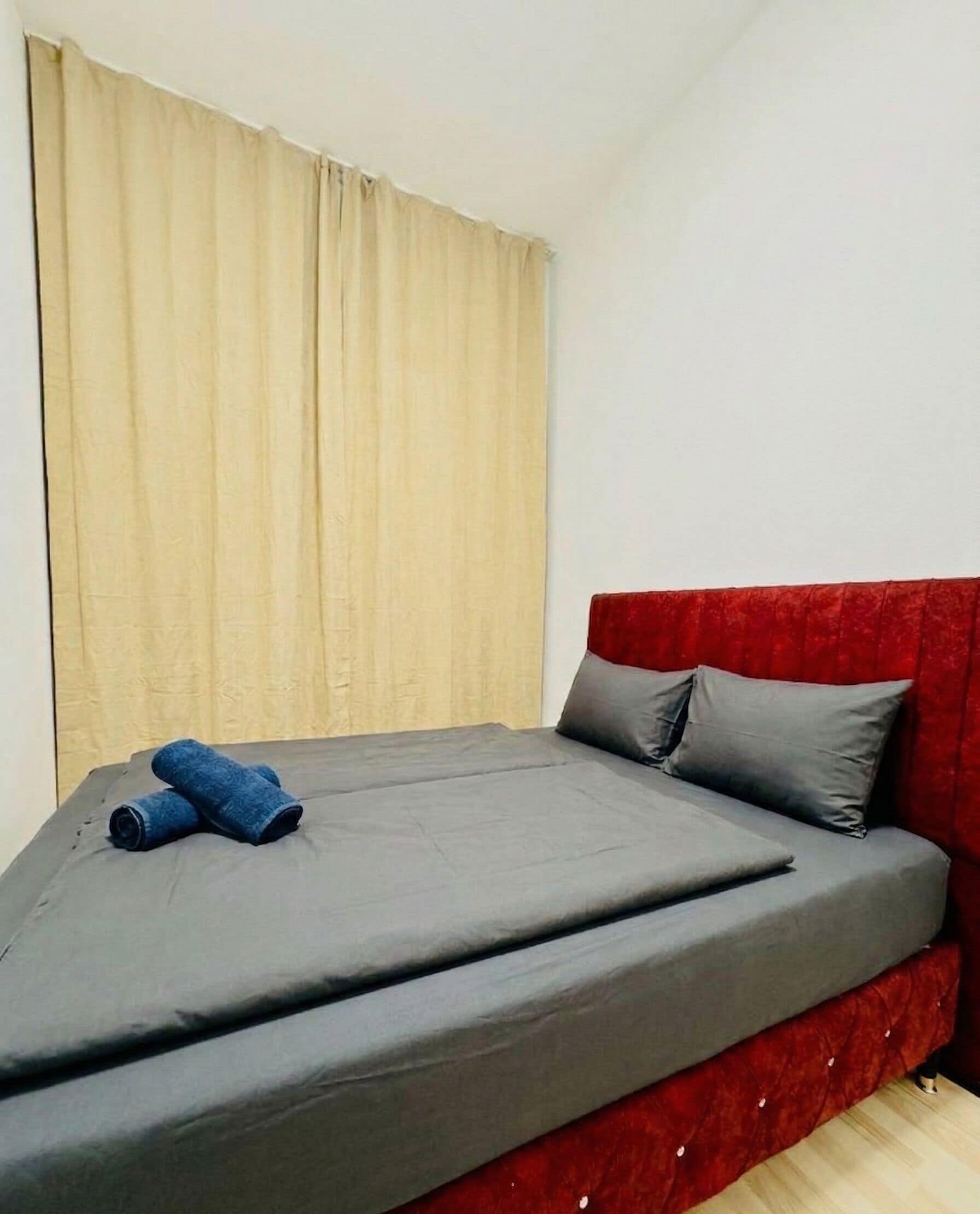 A single bed with a grey mattress and red headboard, a pair of rolled-up blue towels, and beige curtains on a white wall.