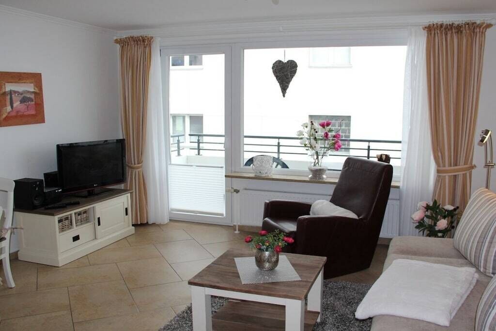 A living room with a television, a sofa, a chair, a small table with a vase, and a balcony with a railing and flowers.