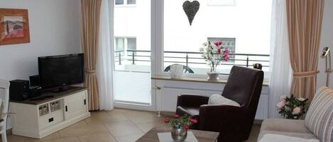 A living room with a television, a sofa, a chair, a small table with a vase, and a balcony with a railing and flowers.