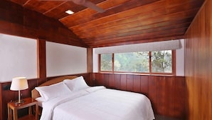 Un dormitorio con techo de madera, una cama con ropa de cama blanca, una mesita de noche con una lámpara y una ventana con vistas a árboles.