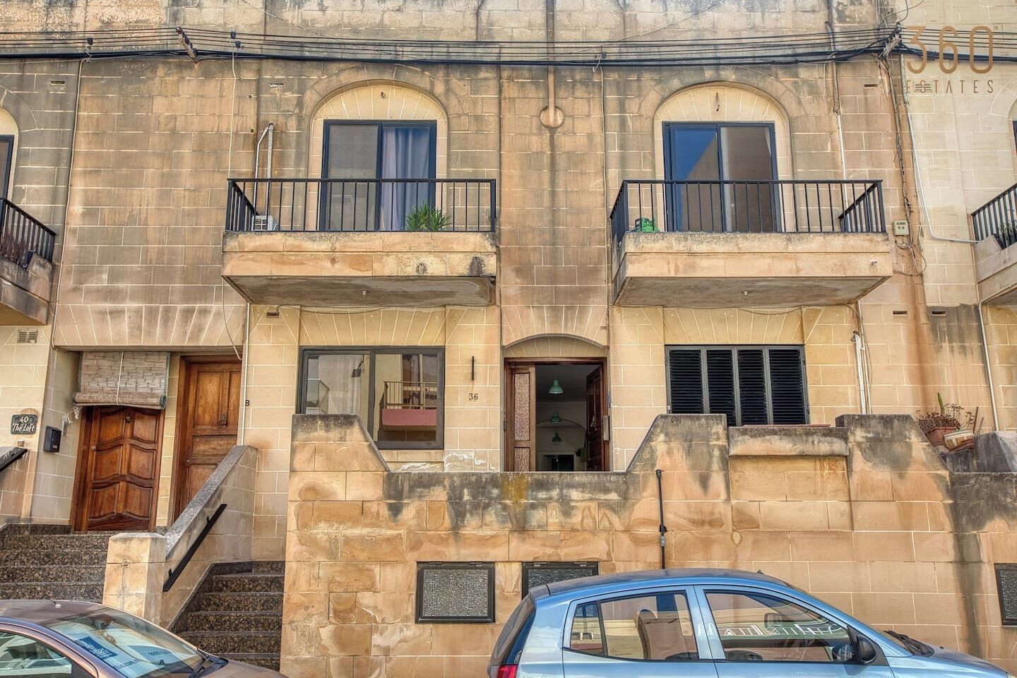 Edificio de dos pisos con fachada de piedra, número 36, que presenta una puerta de madera, un pequeño balcón con barandilla de metal y una escalera que conduce a la entrada.