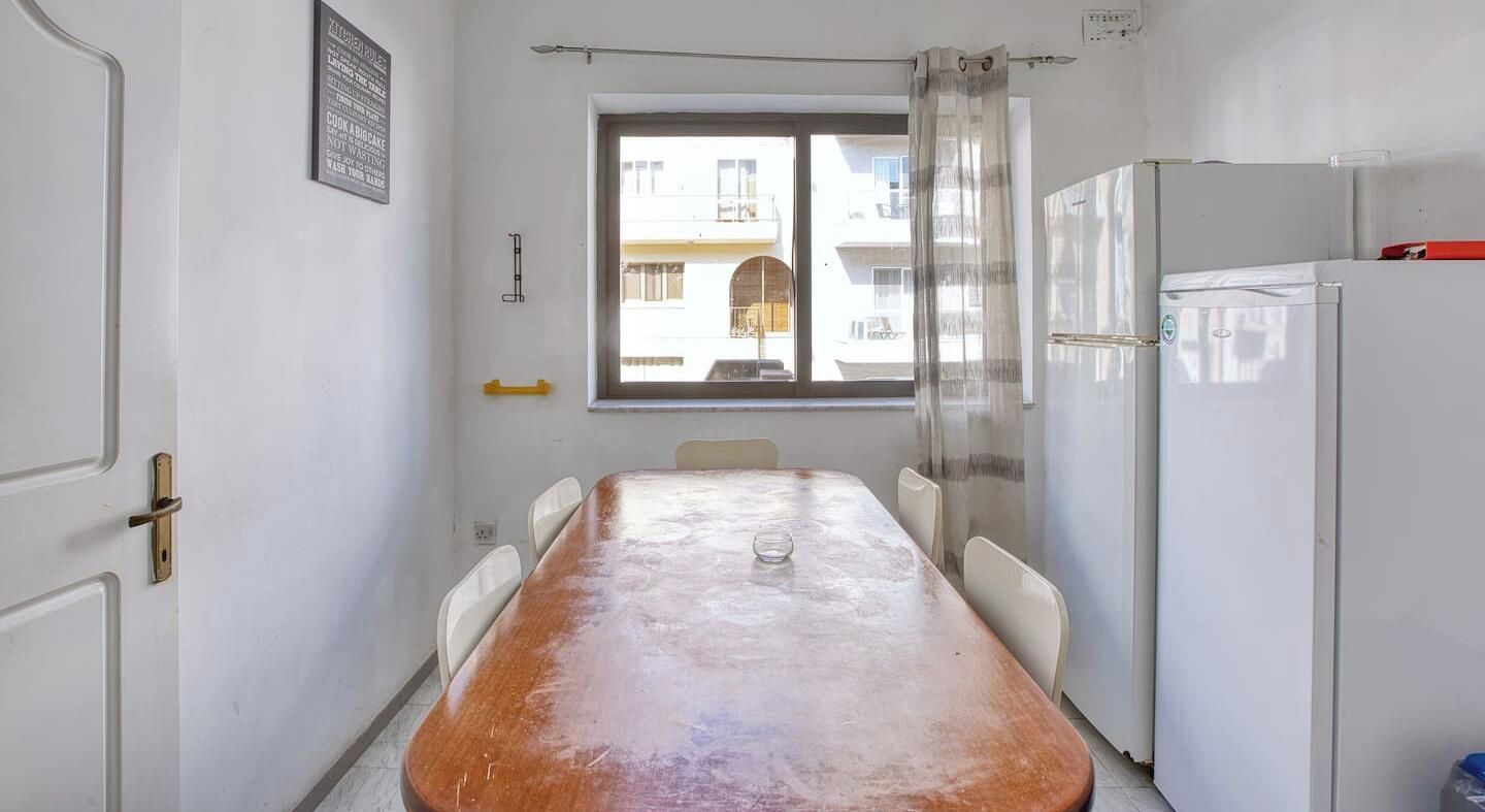 Une petite cuisine avec une table en bois, des chaises blanches, un réfrigérateur et une fenêtre donnant sur les bâtiments à l’extérieur.