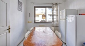 Une petite cuisine avec une table en bois, des chaises blanches, un réfrigérateur et une fenêtre donnant sur les bâtiments à l’extérieur.