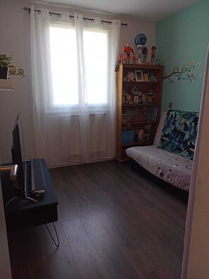 Ein Zimmer mit einem Bett, einem Schreibtisch mit Computer, einem Bücherregal und einem Fenster mit Vorhängen.