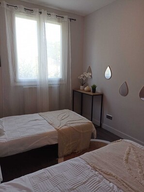 Ein Schlafzimmer mit zwei Betten, einem Nachttisch mit Spiegel und einem Fenster mit durchsichtigen Vorhängen.