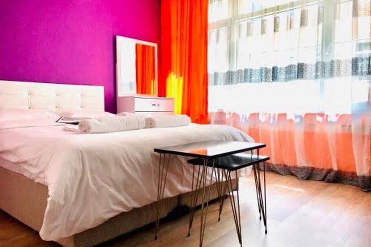 A bedroom with a bed, a bedside table, a mirror, and a window with curtains.