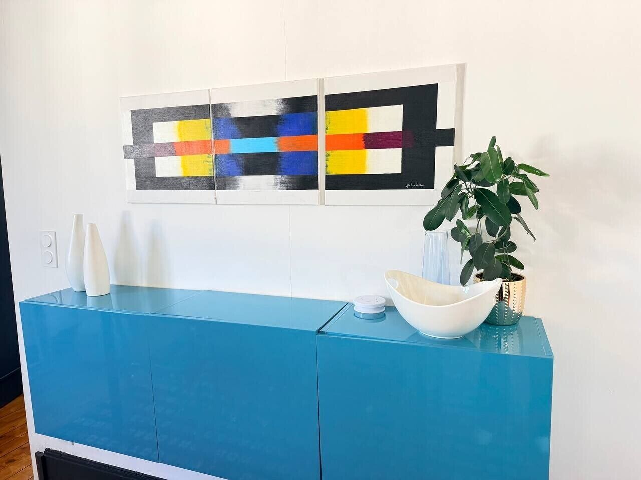 A blue console with a white bowl and vases, a potted plant, and abstract art on the wall.