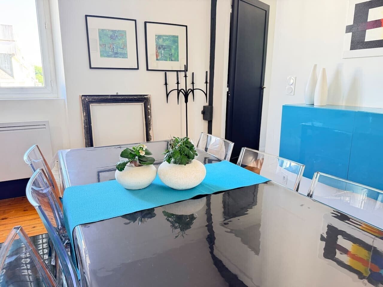 Una mesa de comedor con camino de mesa azul, dos plantas en macetas y sillas transparentes.