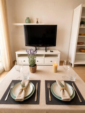 A dining table set with plates, cutlery, and a candle, with a television and storage unit in the background. - Serena Golf Views • Beach & Golf Apartment (Alcazares, Los)