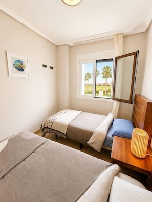 A bedroom with two beds, a wooden nightstand, a lamp, a window with a view of palm trees, and a framed picture on the wall. - Serena Golf Views • Beach & Golf Apartment (Alcazares, Los)