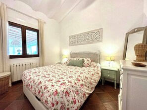 Ein Schlafzimmer mit einem Bett, zwei Nachttischen, einem Heizkörper, einem Fenster mit Vorhängen, einem Wandbild und einem Spiegel mit Dekoration darauf.