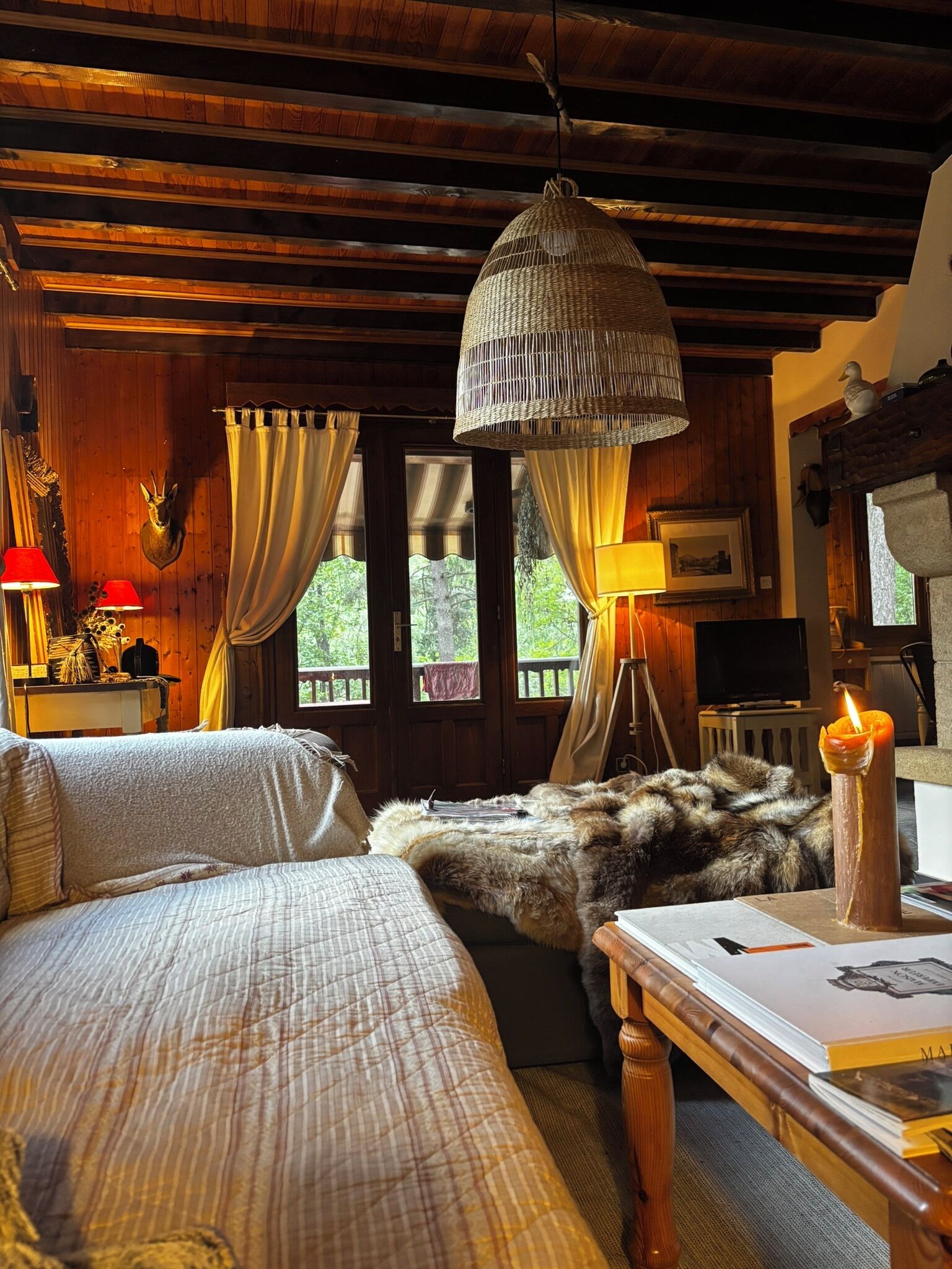 Une pièce chaleureuse avec des poutres apparentes, un lit recouvert d’un plaid en fourrure, une lampe suspendue et une bougie posée sur une table.