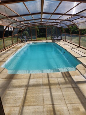 Ein Schwimmbecken mit Glasüberdachung, umgeben von einer Terrasse mit Stühlen.