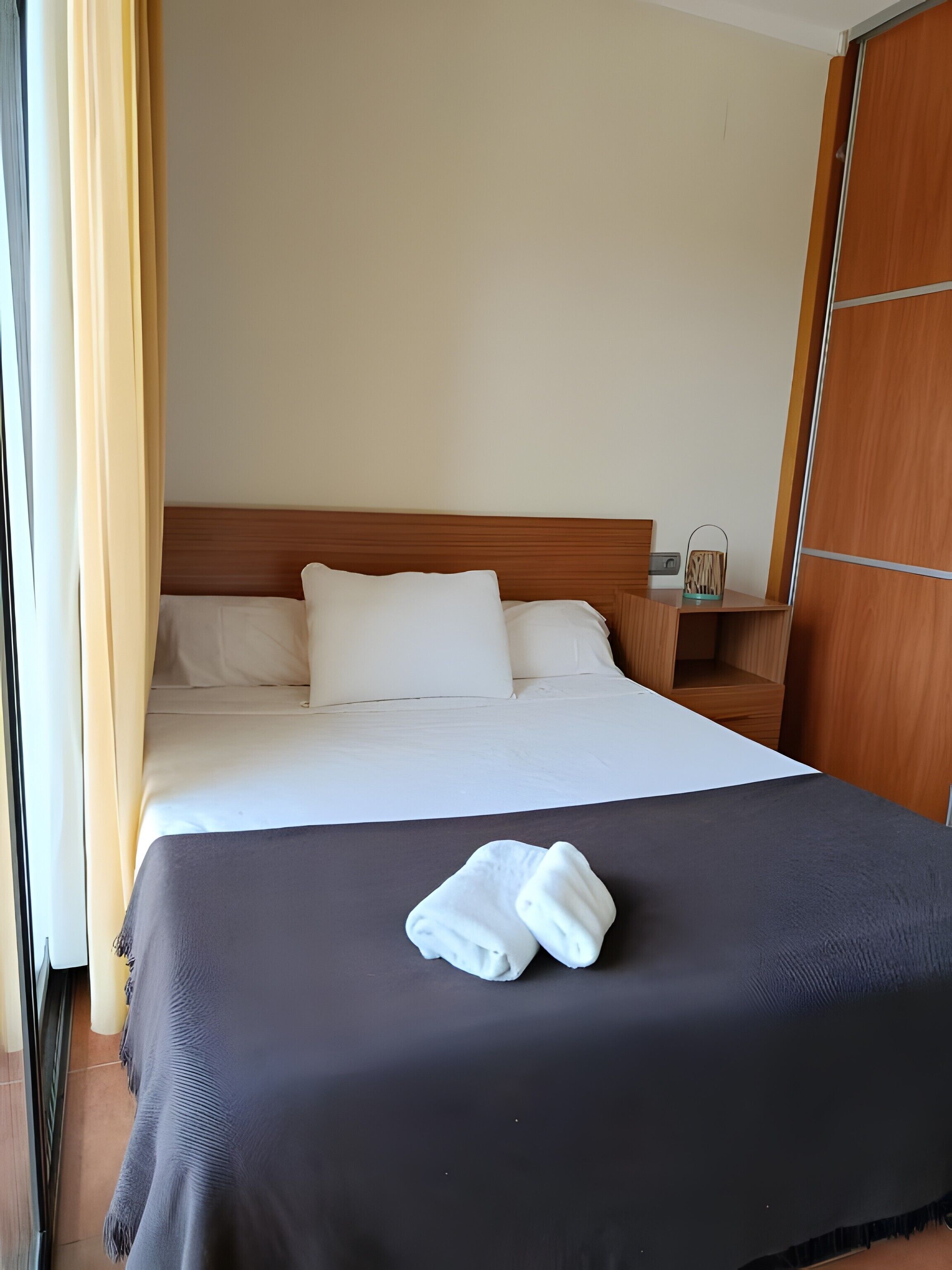 Habitación de hotel con cama, mesita de noche, armario de madera y ventana con cortinas.