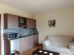 A compact kitchen with wooden cabinets, a microwave, and a sink. There's a beige sofa and a small table with papers.