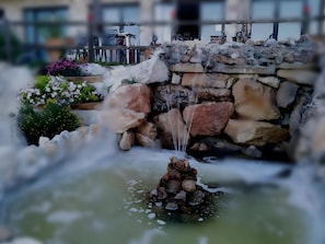 A water feature with a stone structure and a small waterfall, surrounded by greenery and flowers.