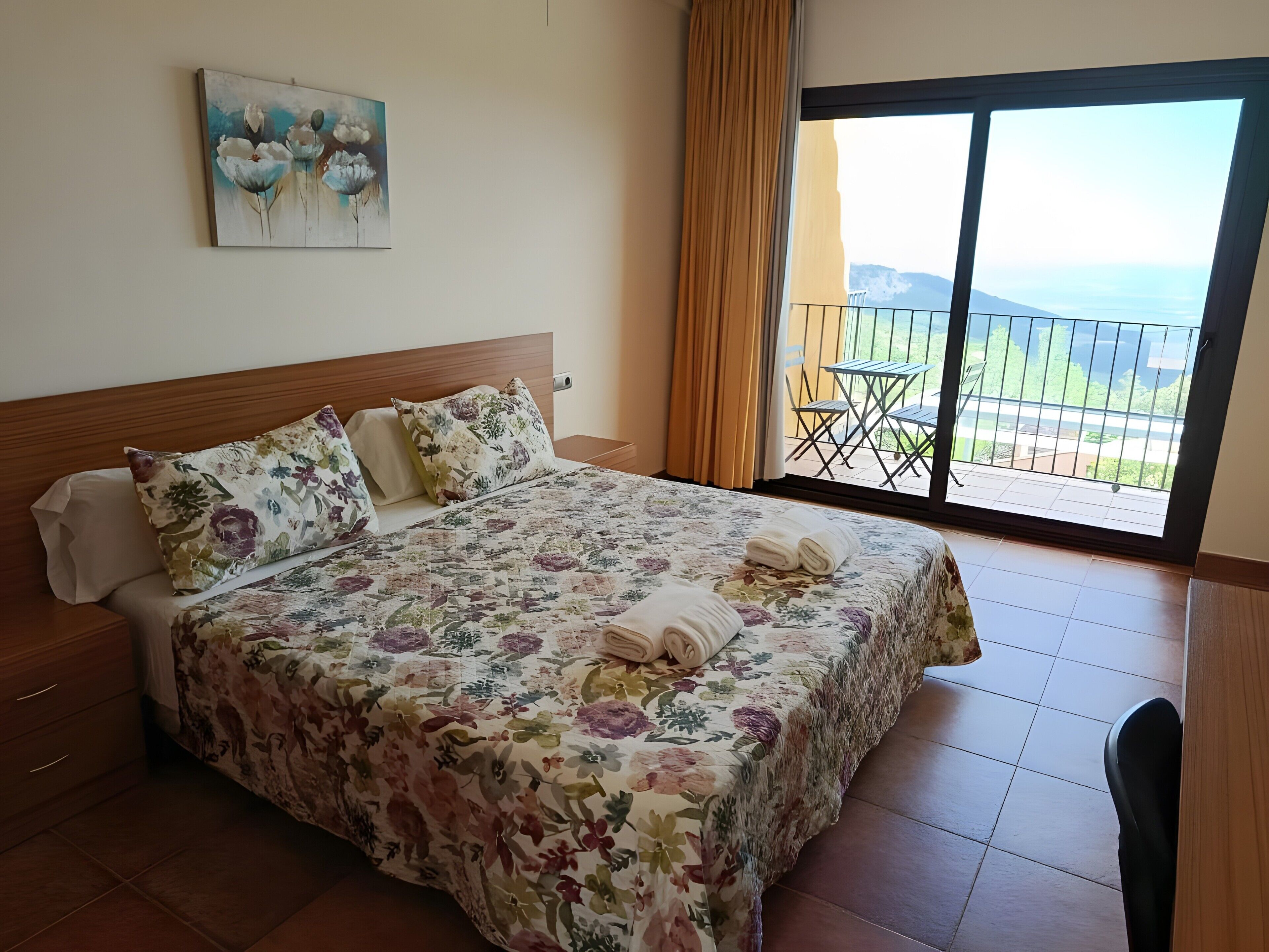 Een hotelkamer met een groot bed, bloemenbeddengoed, een schilderij aan de muur en een balkon met uitzicht.