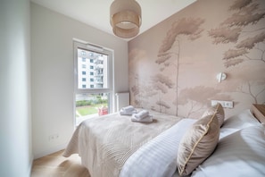 A bedroom with a large window, a bed with pillows, and a wall with a tree mural. - Kierbedzia Apart (Warszawa)