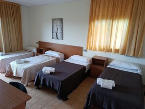 Habitació d'hotel amb quatre llits individuals, un panell de capçalera de fusta, una petita taula de nit de fusta, un quadre penjat a la paret i una finestra amb cortines.