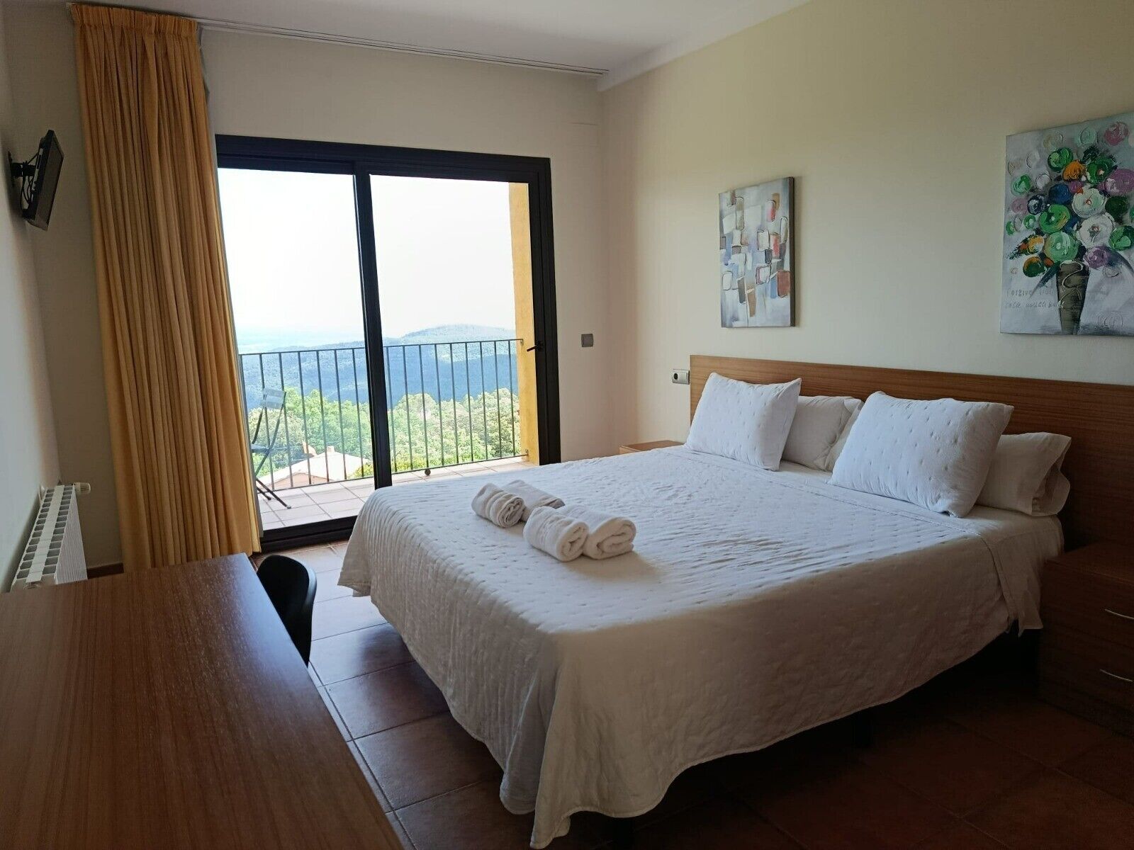 A hotel room with a large bed, a view of the outdoors, a painting on the wall, and a window with curtains.