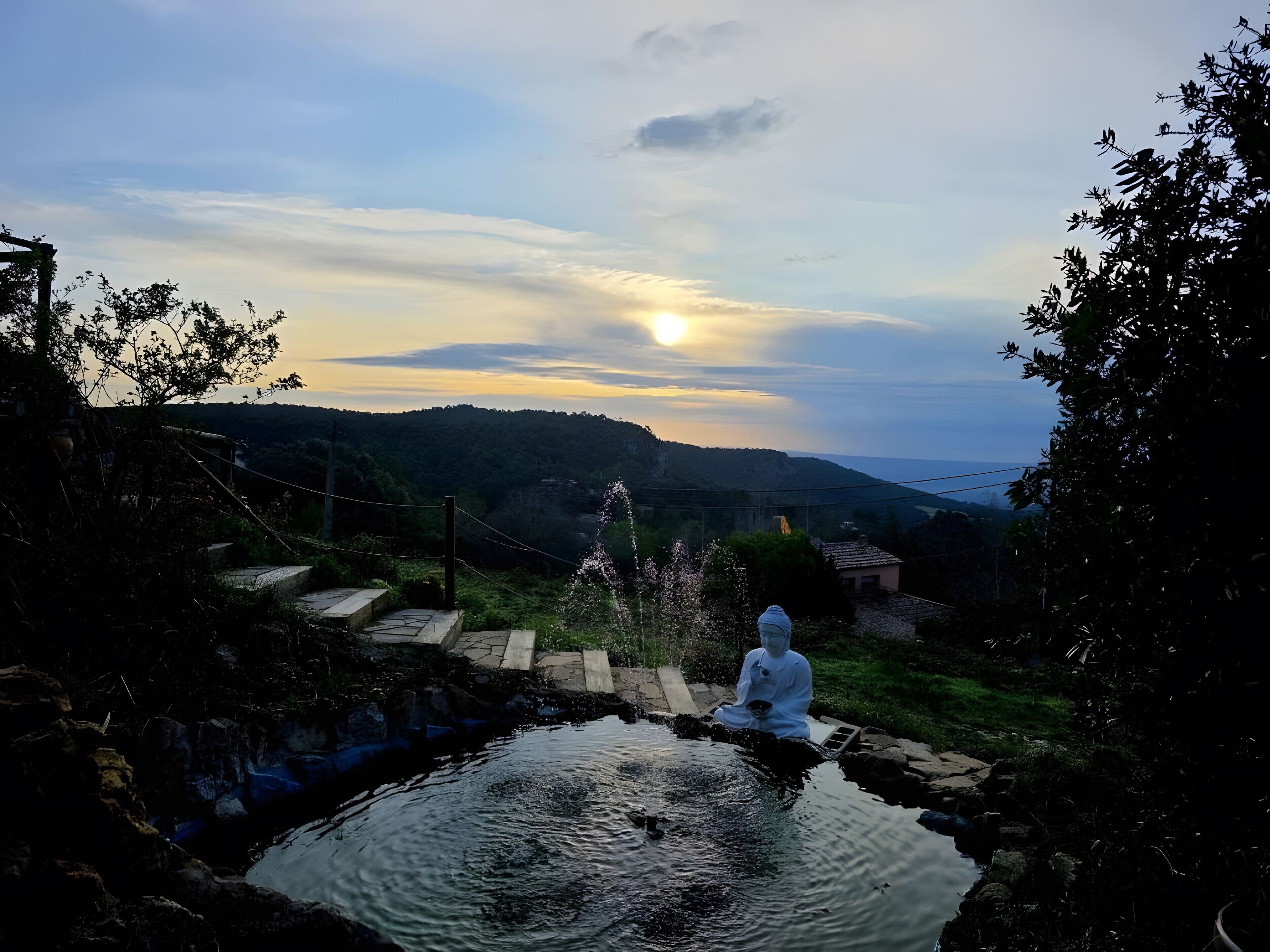 En rogivande utomhusmiljö med en Buddha-staty vid ett stilla vatten, omgiven av grönska och kullar i bakgrunden.