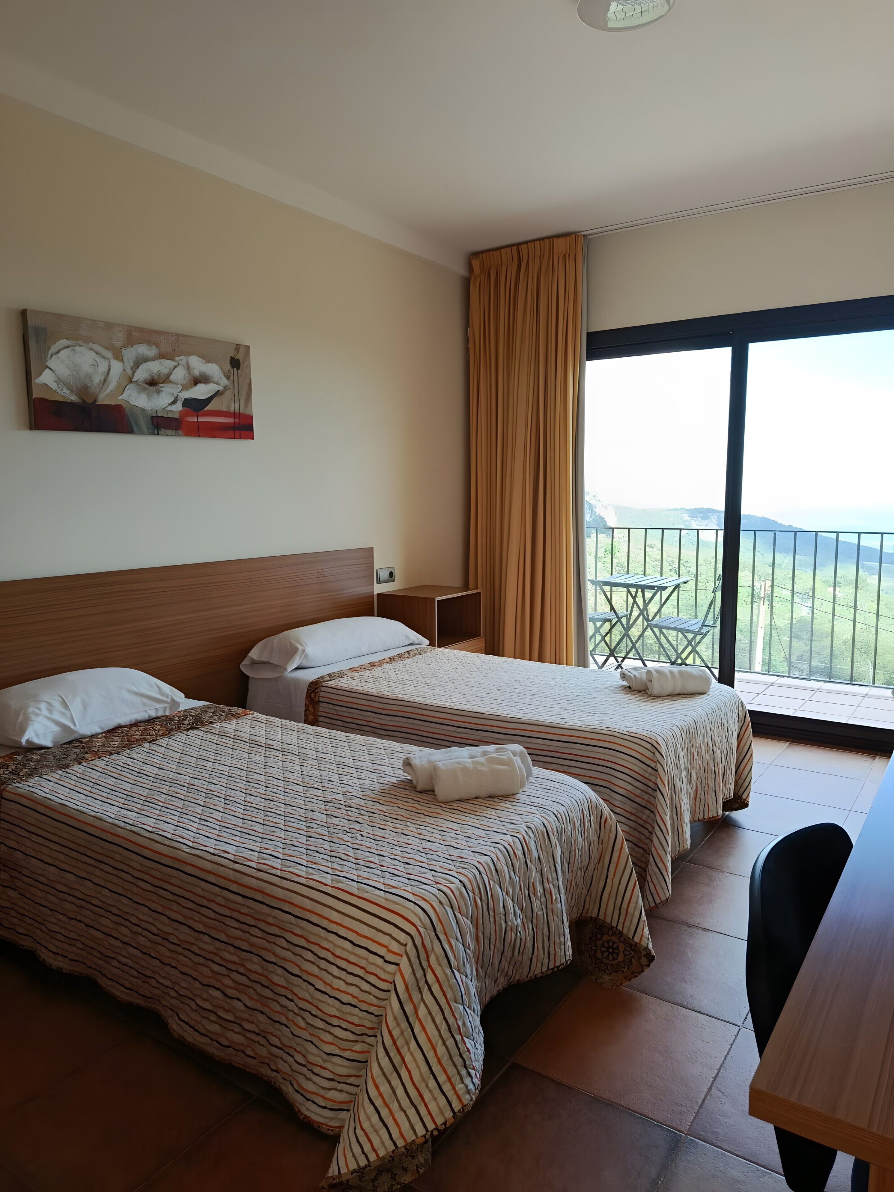 A hotel room with two beds, a painting on the wall, a balcony with a view, and a desk.