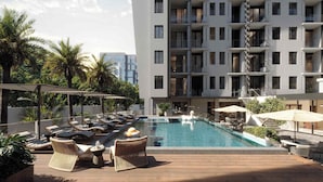 Un área de piscina en la azotea con sillones y sombrillas, rodeada por un edificio moderno con balcones.
