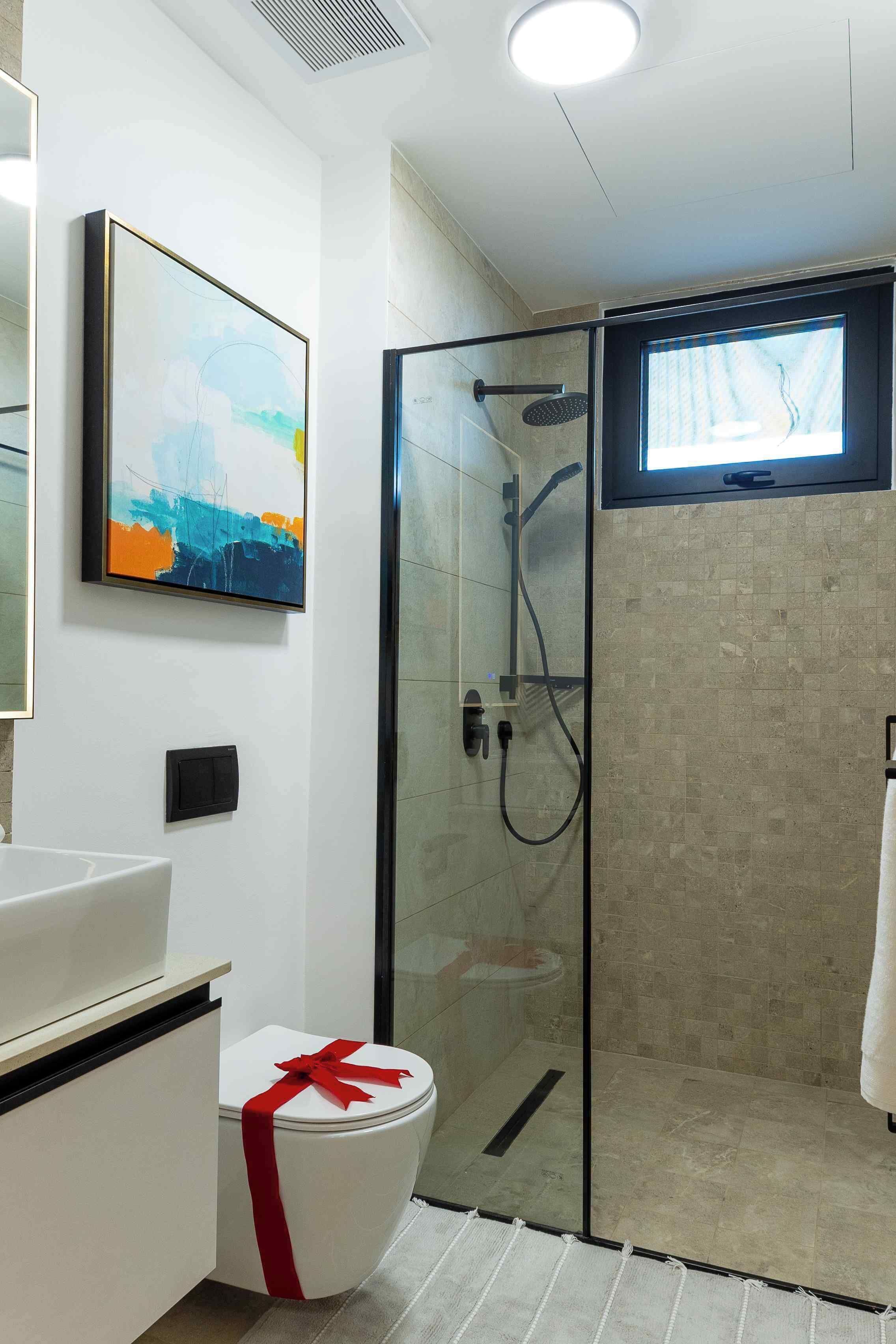 A modern bathroom with a glass-enclosed shower, a white toilet with a red ribbon, and a framed abstract painting on the wall.