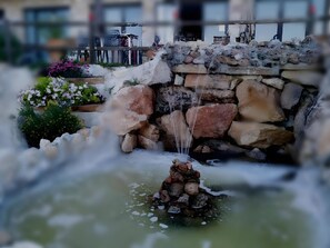 A water feature with a stone structure and a small waterfall, surrounded by greenery and flowers.