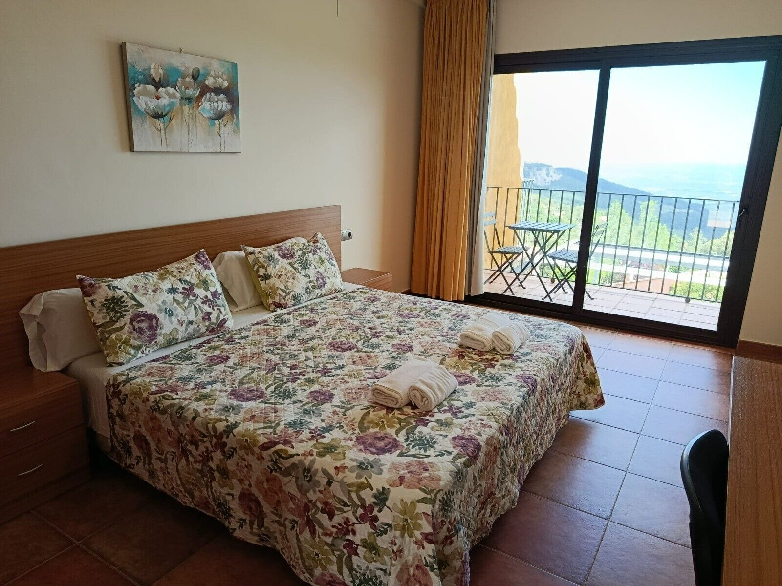 A hotel room with a bed, a painting, a balcony with a view, and a chair.