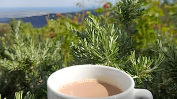 A white cup of tea on a saucer with a tea bag, set against a backdrop of green foliage and a distant view of a landscape.