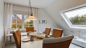 Een eethoek met een witte tafel gedekt voor zes personen, omringd door rieten stoelen, een hangende lamp en een raam met uitzicht op een huis en bomen.