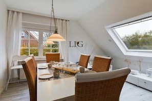 A dining area with a white table set for six, surrounded by wicker chairs, a hanging light fixture, and a window with a view of a house and trees. - Holiday apartment Seestern (Langeoog)