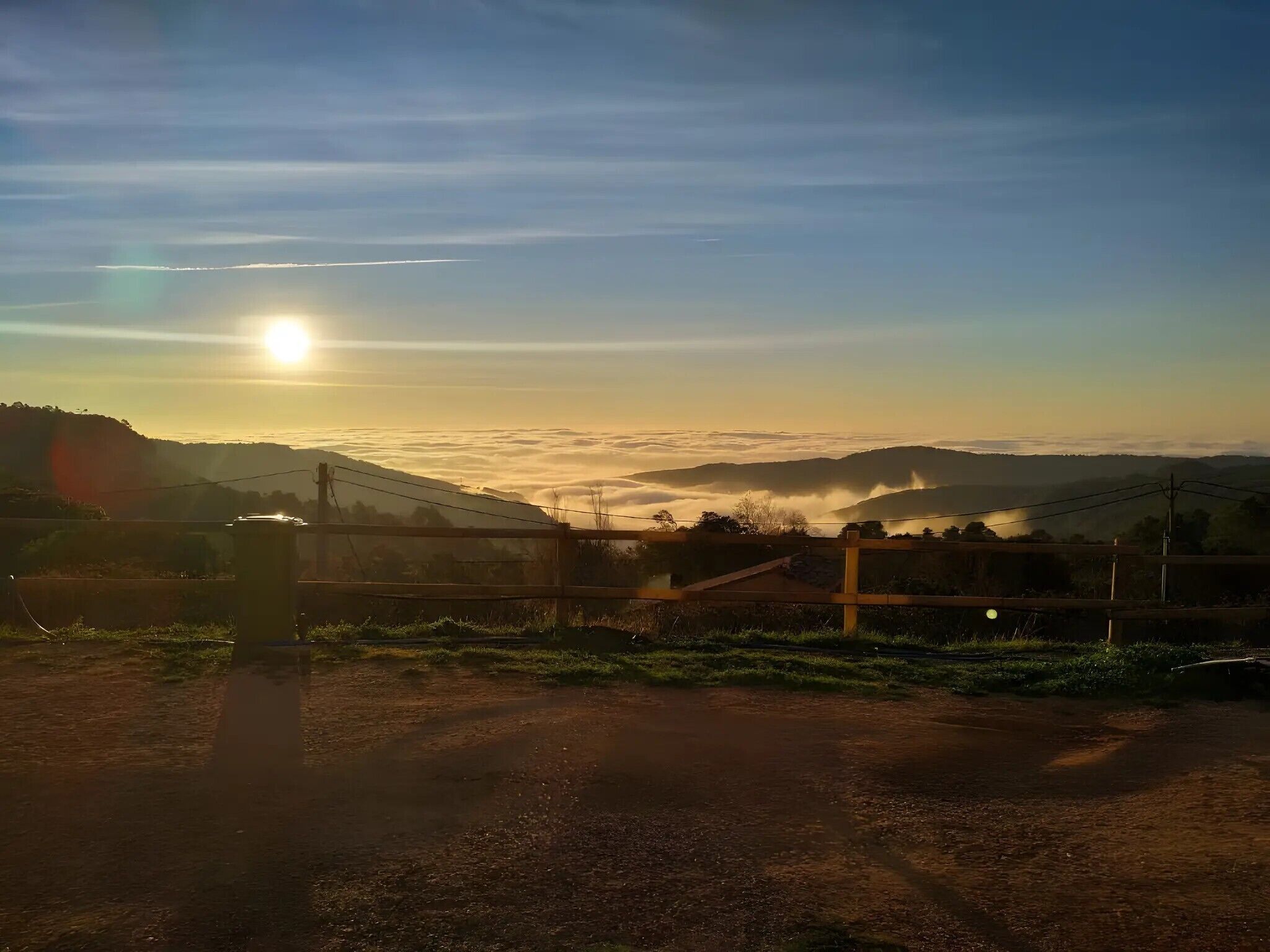 Landskab med åben himmel, lavstående sol og et hegn i forgrunden.