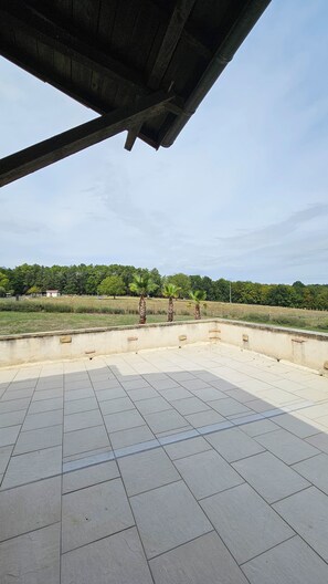 An outdoor area with a tiled floor, a concrete structure overhead, and a view of a green landscape with trees and a fence in the background. - Vrbo Property (Agonac)