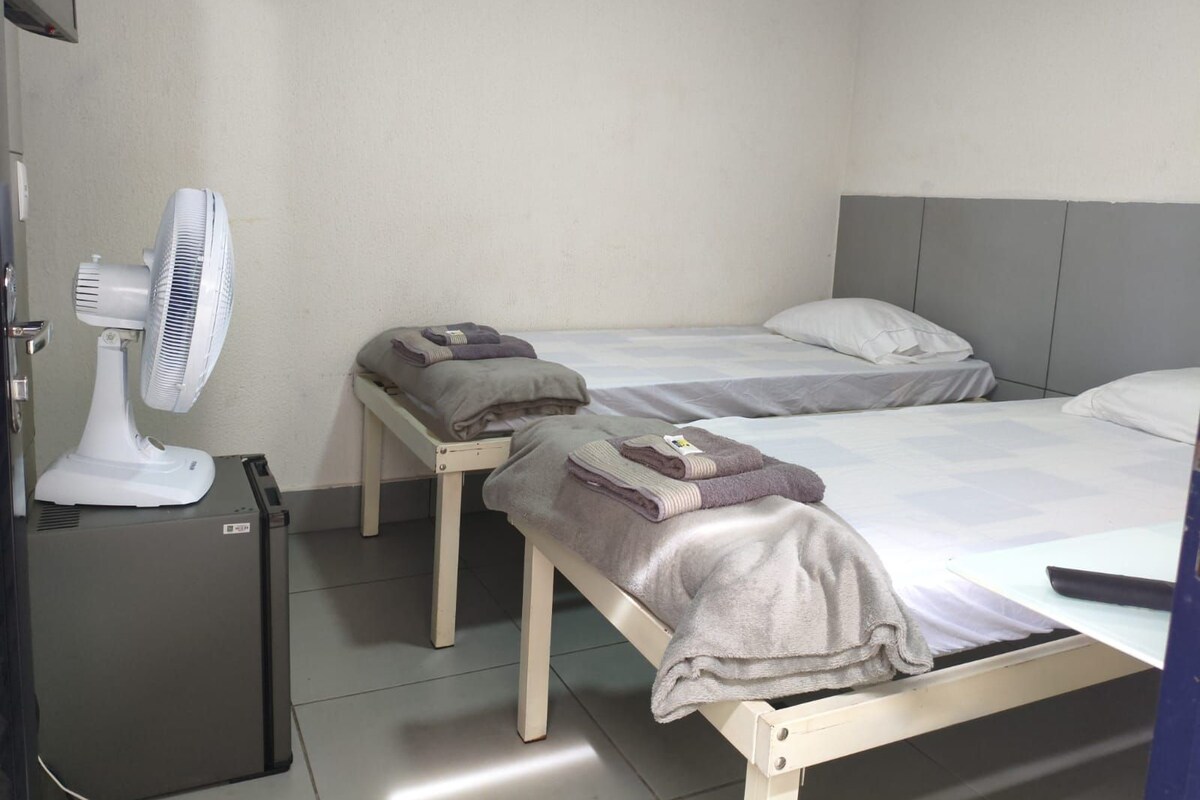 A hotel room with two single beds, a bedside table with a towel, a fan, and a small refrigerator.