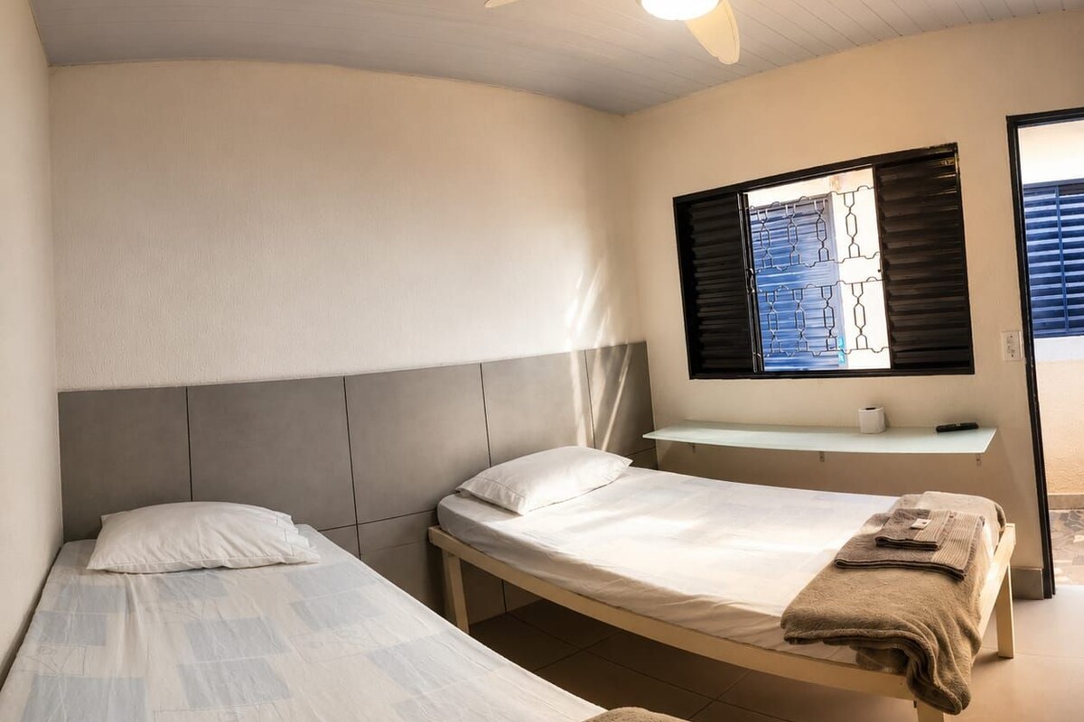 A bedroom with two single beds, a ceiling fan, a window with blinds, and a bedside table with a towel.