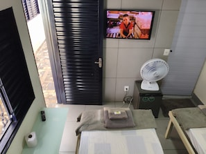 A hotel room with a bed, a TV mounted on the wall, a fan on a small table, and a window with blinds.