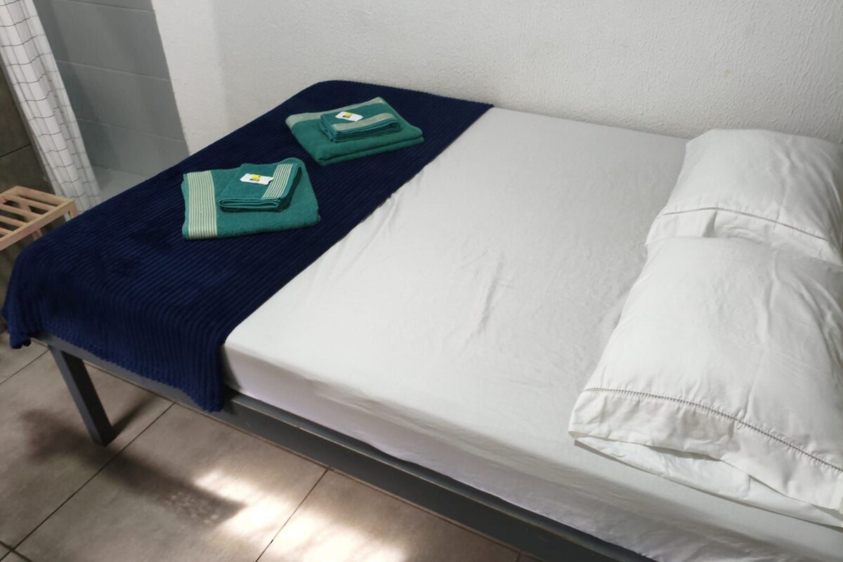 A single bed with a white mattress and pillow, a blue blanket, and two folded green towels.