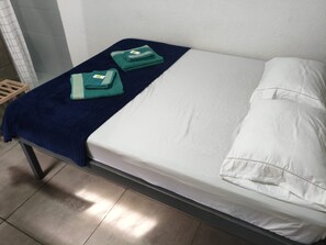 A single bed with a white mattress and pillow, a blue blanket, and two folded green towels.