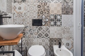 Een badkamer met een witte wastafel, toilet en een douchetoilet. De muren zijn betegeld met een patroon.