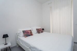 Een eenpersoonsbed met wit beddengoed, twee kussens, een nachtkastje met een lamp, en een raam met dunne gordijnen.