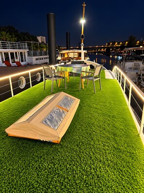 En udendørs siddeplads med et bord og stole på en græsdækket terrasse med udsigt over en lysthavn med både.