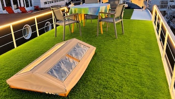 Una terraza en la azotea con césped artificial, una mesa con sillas y vistas a barcos y un puente al fondo.