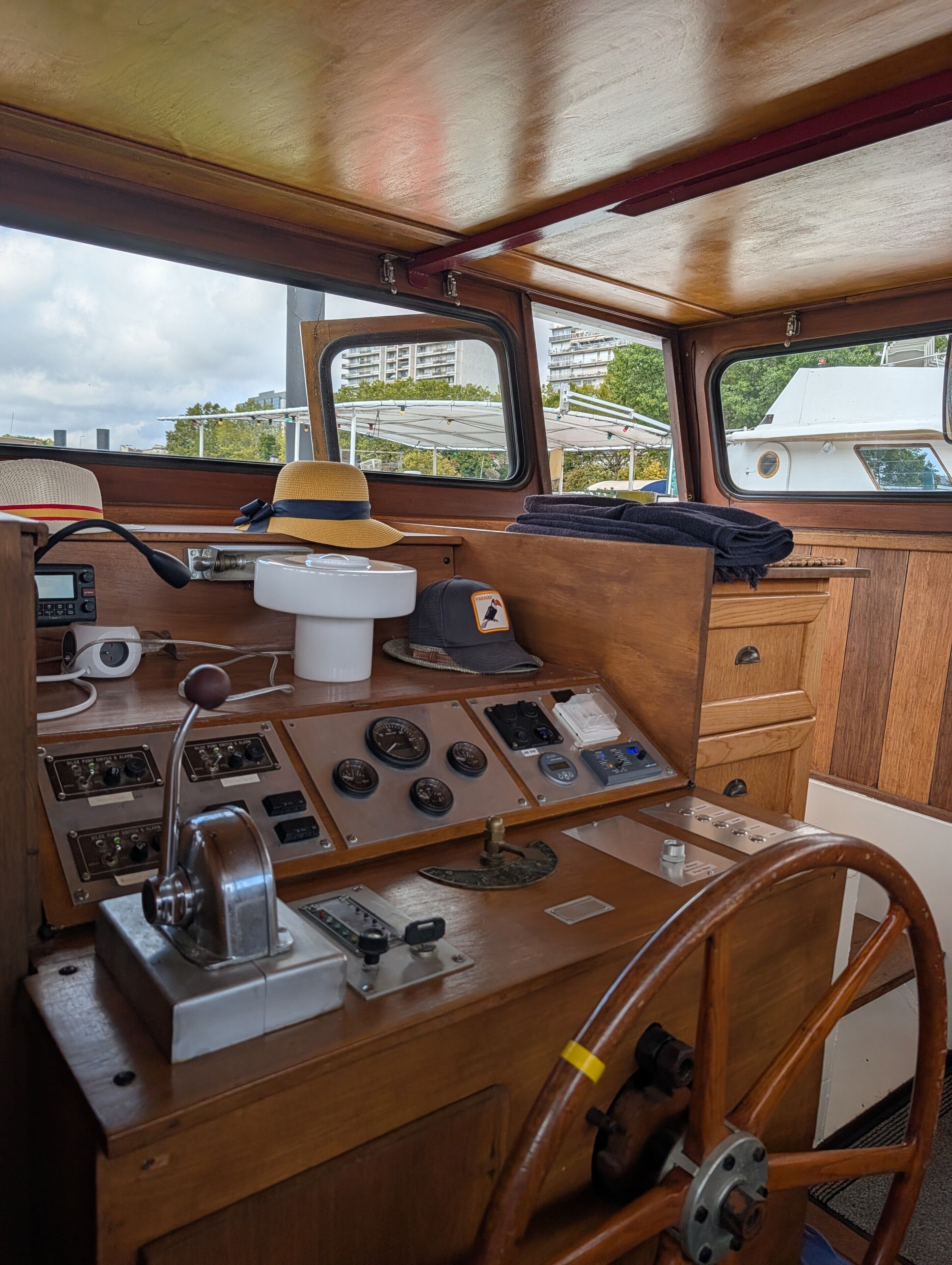 Un poste de pilotage de bateau, doté de divers cadrans, d’un volant et d’une fenêtre offrant une vue sur les bâtiments et les arbres.