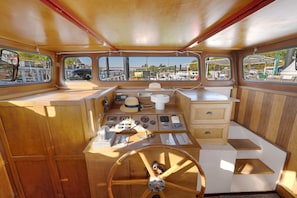 A boat's interior with a steering wheel, control panels, and windows offering views of a marina.