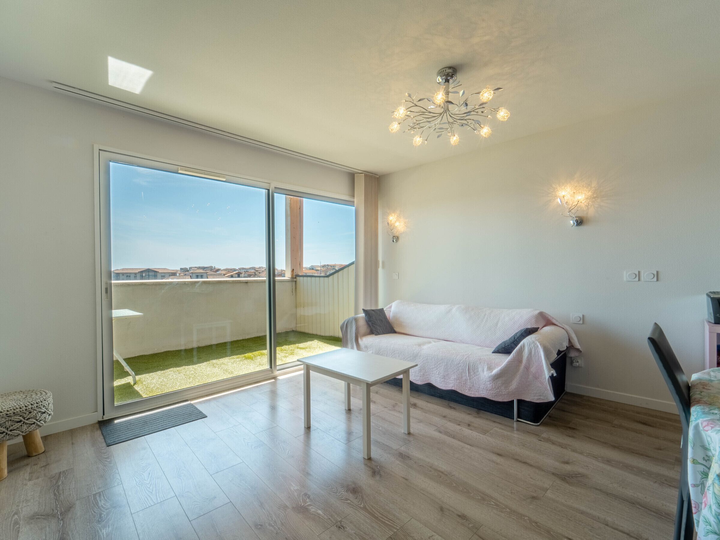 Ein modernes Wohnzimmer mit einer Glasschiebetür, die auf einen Balkon führt, einem weißen Sofa, einem kleinen Tisch und einer Chandelüster.