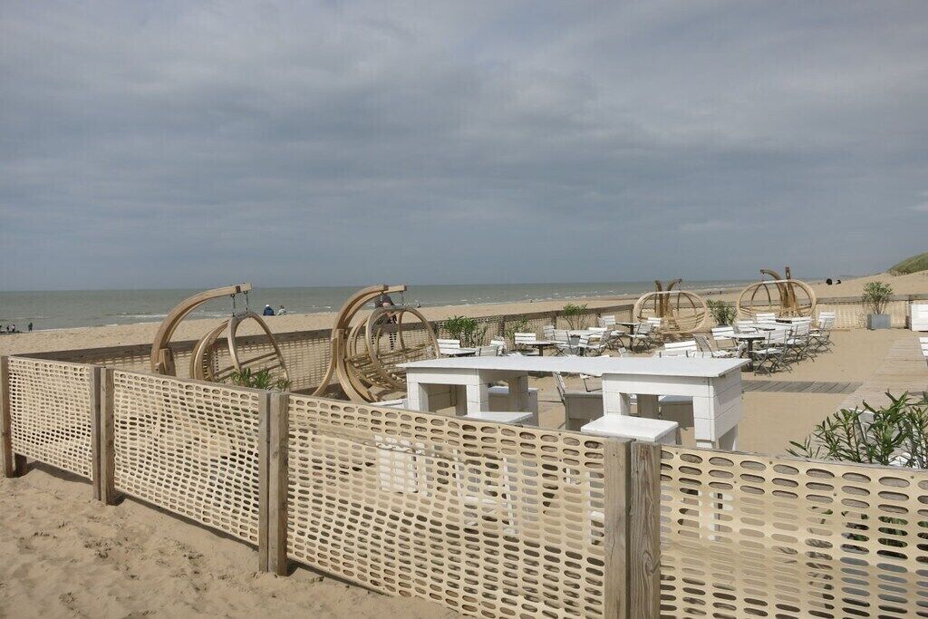 A beachside seating area with tables and chairs, a wooden fence, and a sandy beach.